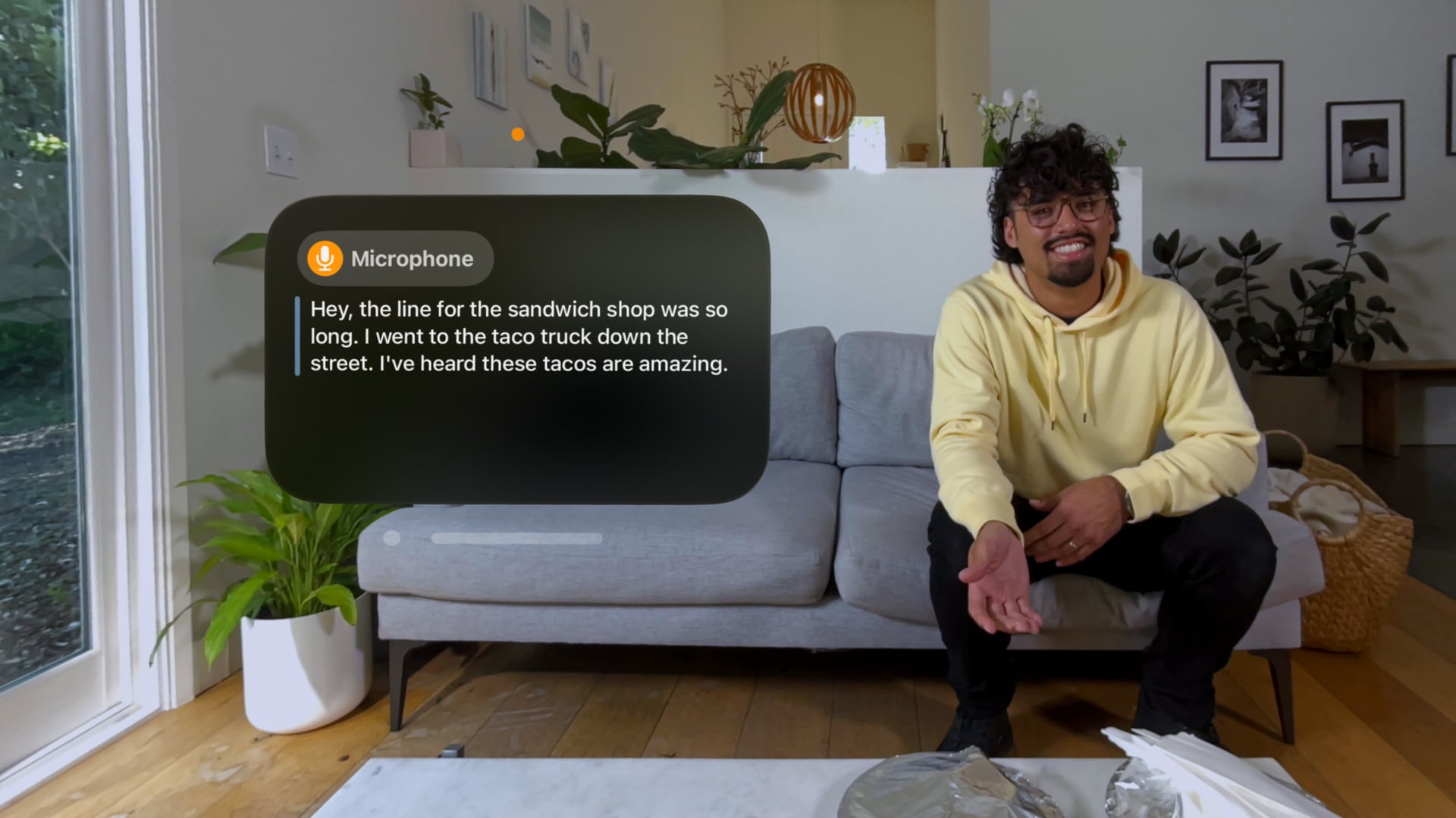 A scene from the Vision Pro headset showing a young man sitting on a couch in a modern apartment, with a live captions bubble showing transcript of their speech in real time