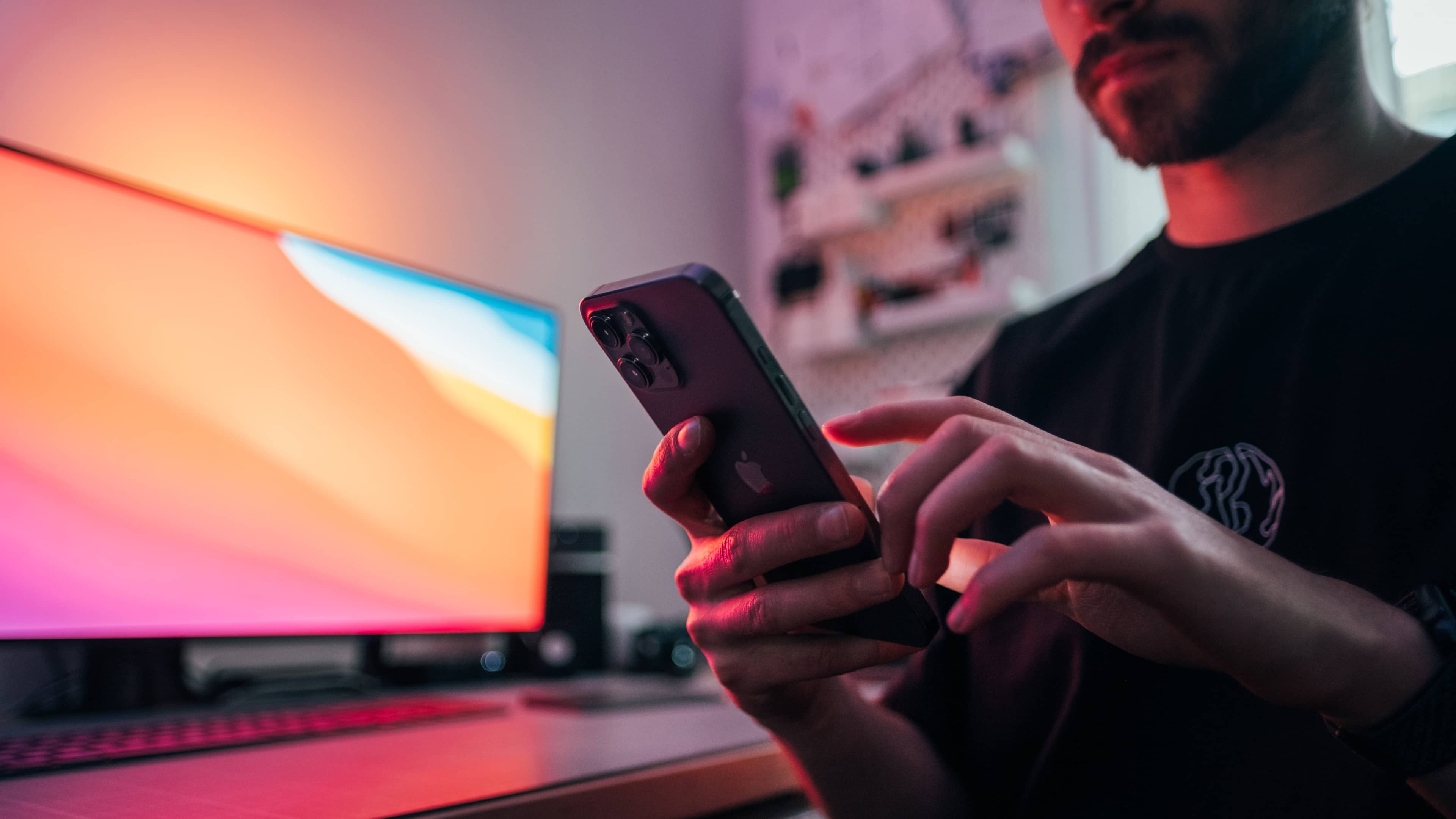 Young man holding an iPhone 12 Pro Max in front of his work desk. In the background, there's a monitor using the Big Sur wallpaper and Apple's space gray keyboard and trackpad