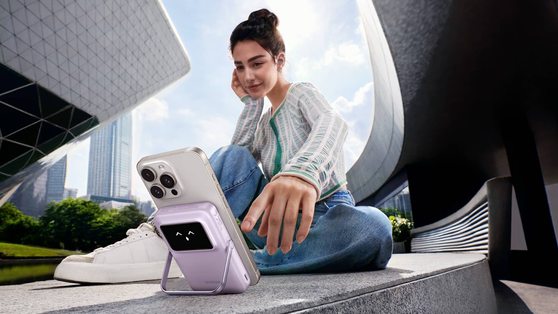 Young woman sitting in an urban environment, looking at her iPhone with a Ugreen powerbank snapped on its back and used as a stand.