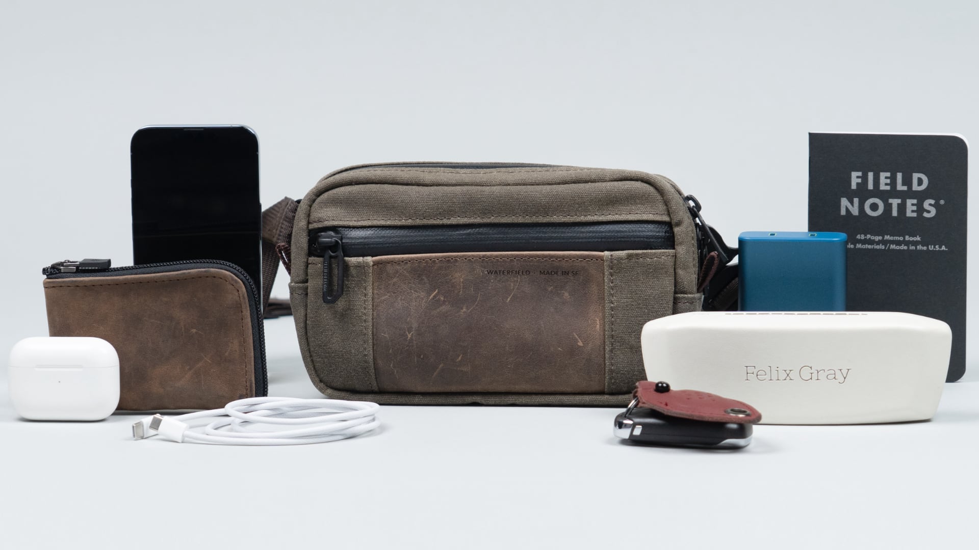 WaterField's Shinjuku Sling crossbody tech case, surrounded by various items that fit inside.