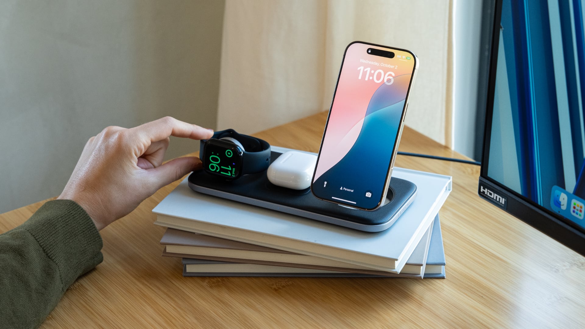 Satechi's Qi2 wireless charger with iPhone, Appel Watch and AirPods on it, resting on a pile of books atop a wooden desk.