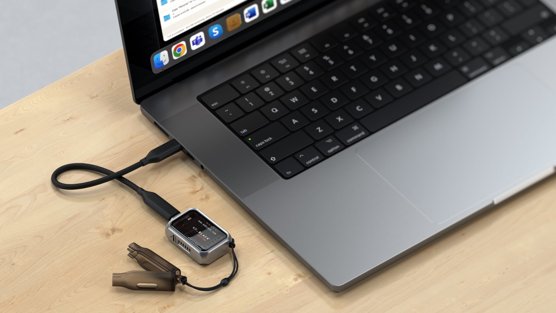 Top-down view of a MacBook Pro on a wooden desk with a Satechi SSD enclosure plugged into its USB-C port.